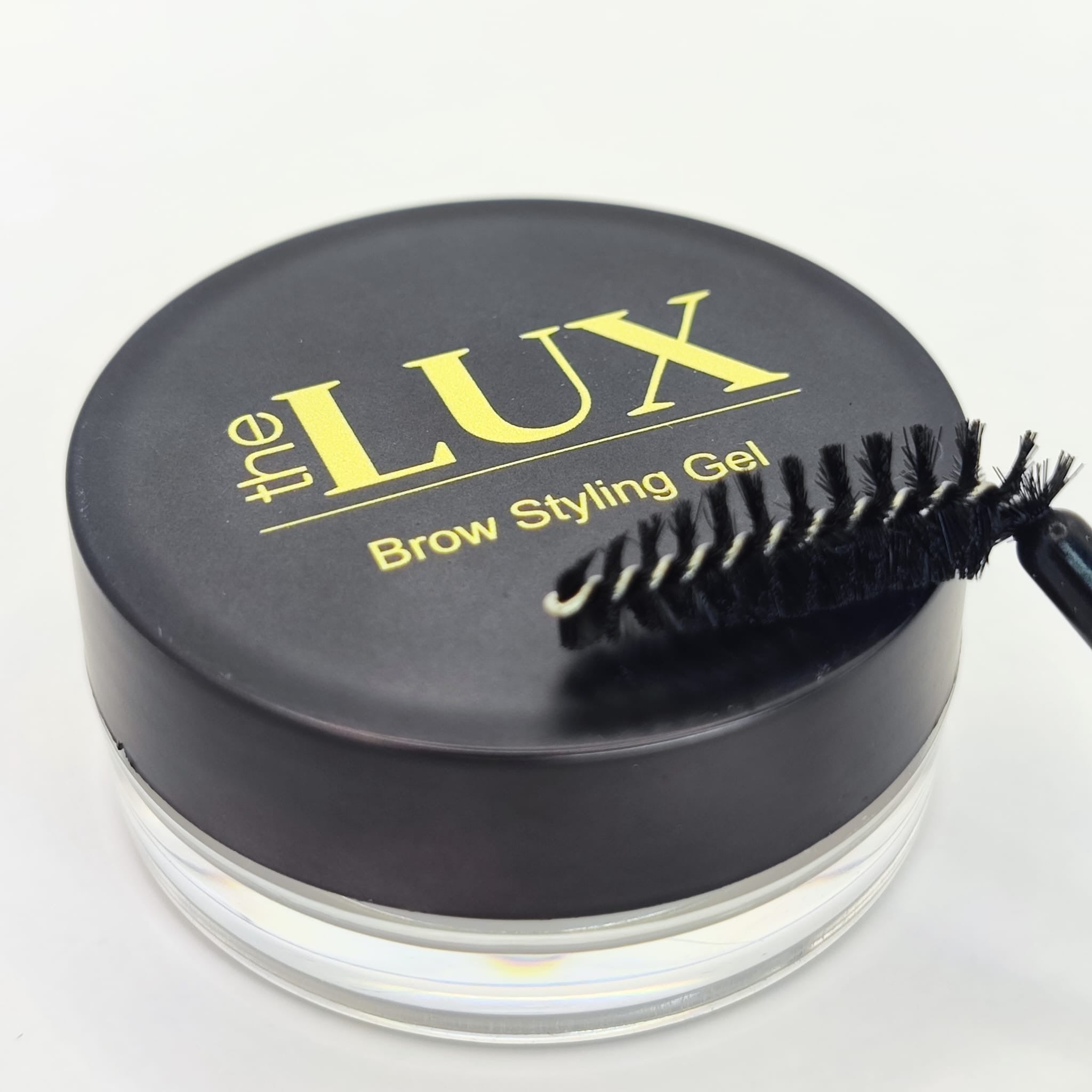 The Luxe Brow Styling Gel black circular tin and retail box shown with a professional tapered spoolie brush for precision brow shaping and setting.